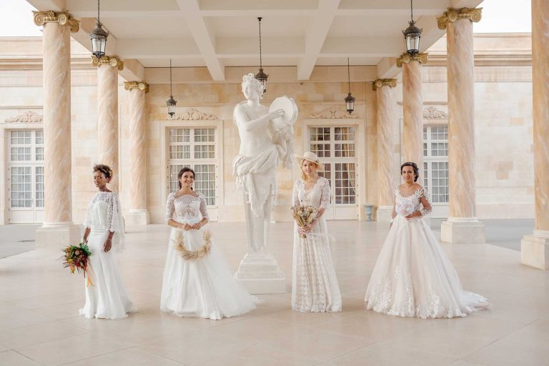 Quatre mariées en robes élégantes posant devant une statue dans un cadre architectural somptueux, illustrant la diversité et l'élégance des créations de 'C'est Maman Qui La Fée'