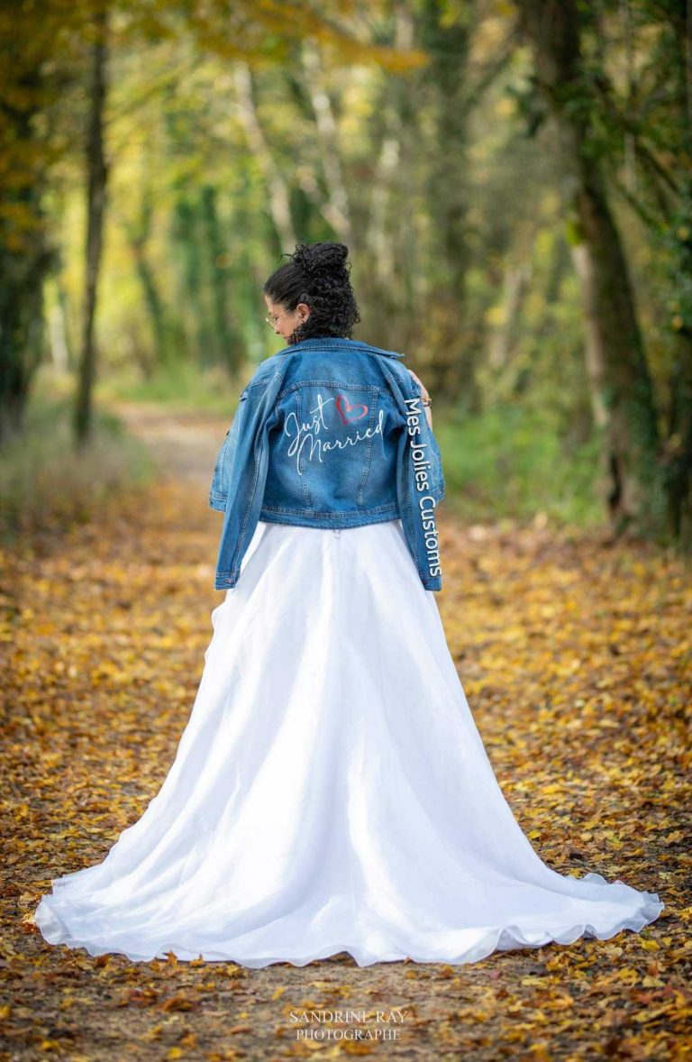 Mariée de dos portant une veste en jean 'Just Married' sur une robe blanche longue, en automne.