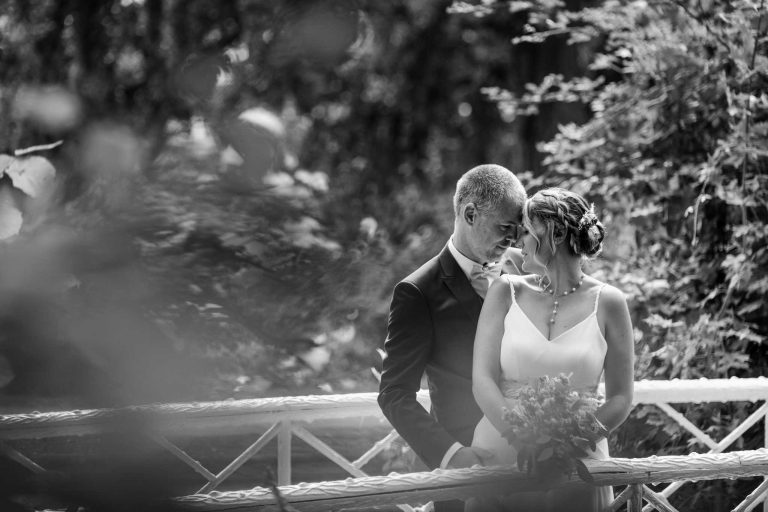 Mariés échangeant un regard tendre sur un pont, la mariée tenant un bouquet, en noir et blanc.