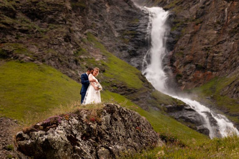 Couple de mariés se tenant devant une cascade majestueuse dans un cadre montagneux