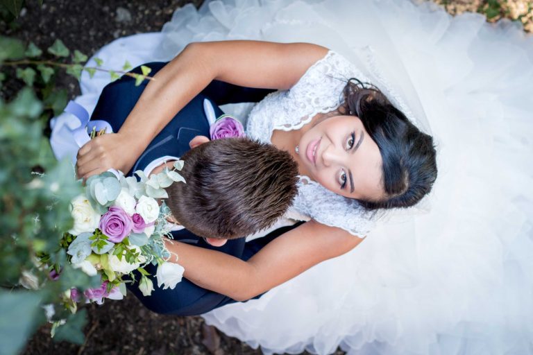 Un couple de mariés vu de dessus, la mariée souriante en robe blanche ornée de dentelle et le marié en costume bleu, tenant un bouquet avec des roses violettes et blanches.