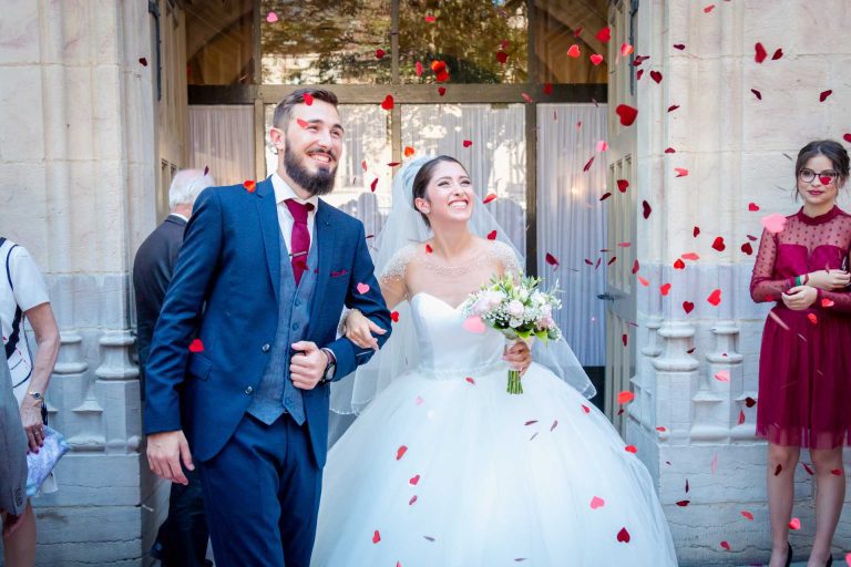 Couple joyeux sortant d'une église sous une pluie de pétales de roses rouges avec des invités en arrière-plan.