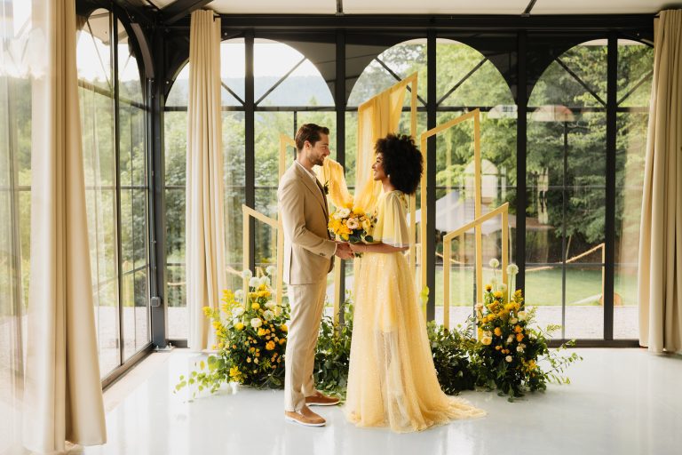 Photo d'un couple dans une piéce avec une femme habiller en robe jaune et un homme qui porte un costume marron claire et qui s'observe autours ont peut voir de grande fenetre ou ont observe le jardin et sur le cote du couple ont peut observer un bouquet de fleurs