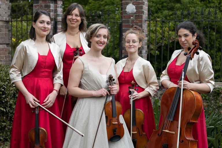 Quatuor à cordes féminin en robes rouges et veste crème posant avec leurs instruments dans un jardin.