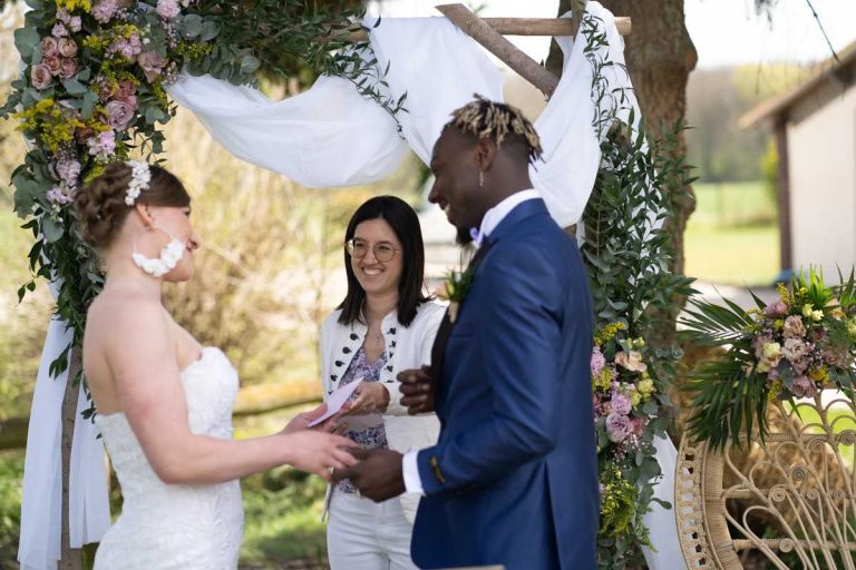 Mariés échangeant leurs vœux en présence d'une officiante, sous un arc floral.