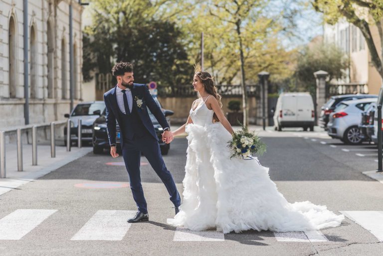 Mariés main dans la main traversant un passage piéton en ville, la mariée en robe blanche volumineuse et le marié en costume bleu.