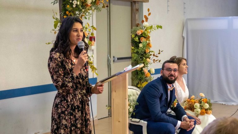 Officiante prononçant un discours lors d'une cérémonie de mariage, avec les mariés assis en arrière-plan.