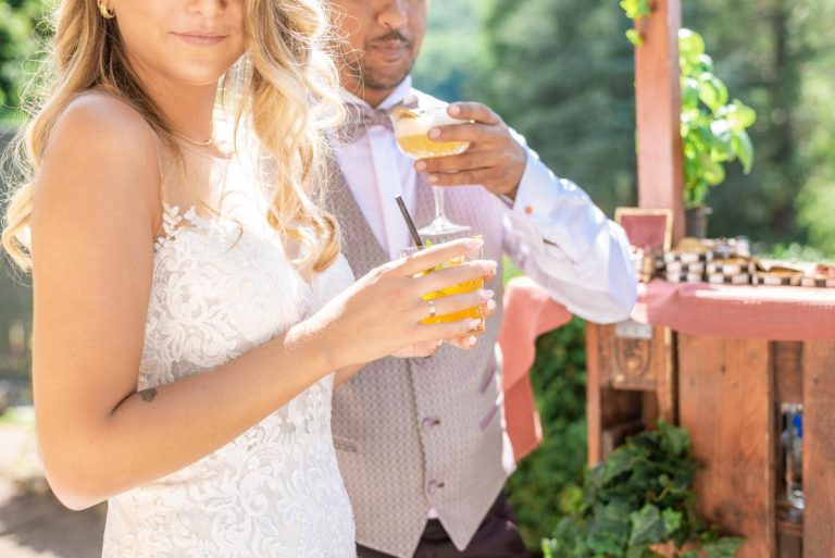 Mariée tenant un cocktail avec le marié en arrière-plan lors d'une réception de mariage en plein air.
