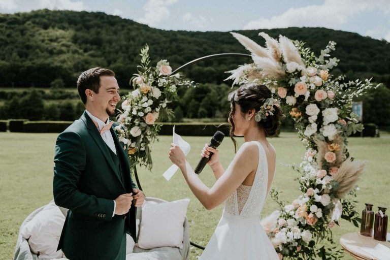 Mariée lisant ses vœux au marié devant un arc floral en plein air.