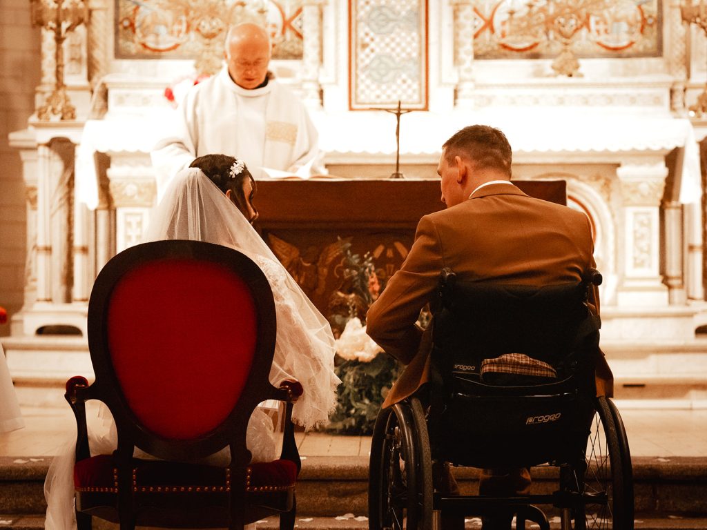 Mariée assise à côté du marié en fauteuil roulant à l'église, écoutant le prêtre pendant la cérémonie de mariage.
