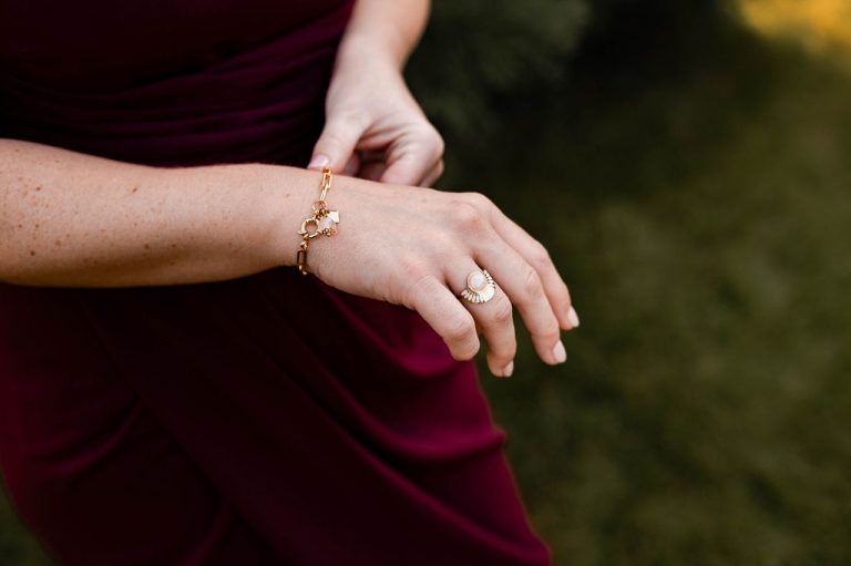 Main de femme avec un bijou sophistiqué sur un bracelet, portant une bague assortie, sur fond de robe bordeaux.