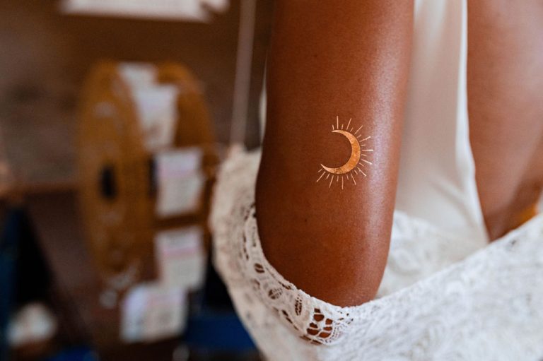 Gros plan sur un tatouage doré en forme de soleil sur l'épaule d'une femme portant une robe blanche en dentelle.