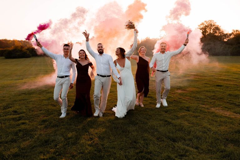 Groupe de mariés et d'amis levant les bras en l'air avec des fumigènes colorés dans un champ au coucher du soleil.