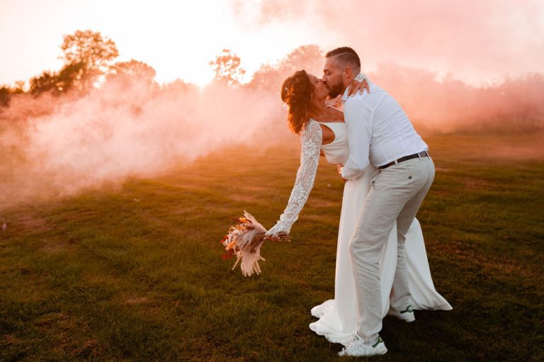 Mariés s'embrassant passionnément sous un ciel coloré par de la fumée rose.