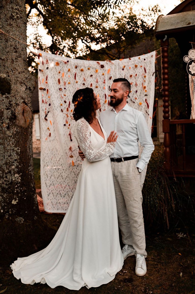 Couple marié se tenant la main devant un rideau bohème orné de fleurs séchées.