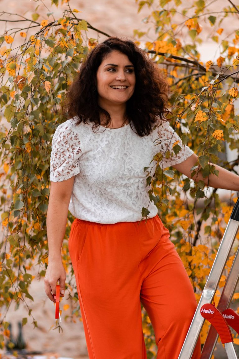 Femme souriante en blouse en dentelle blanche et pantalon orange tenant une échelle près d'un arbre.