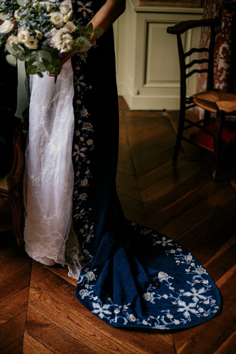 Détail d'une robe de mariée avec traîne bleue ornée tenant un bouquet, dans un intérieur élégant.