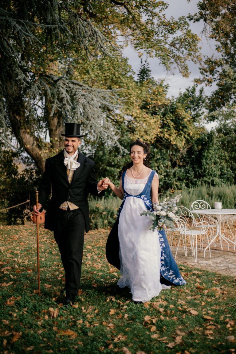 Mariés souriants marchant ensemble en extérieur, le marié portant un haut-de-forme et une canne.