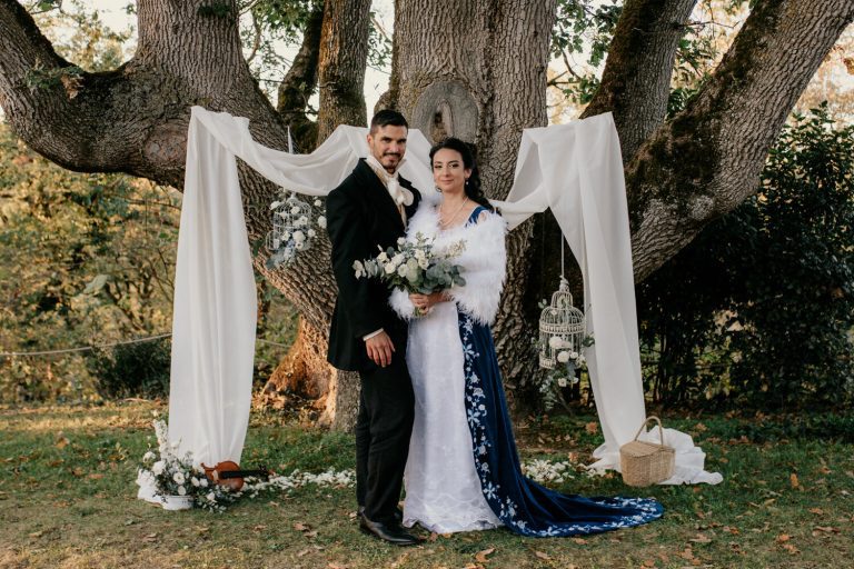 Couple marié debout sous un grand arbre orné de drapés blancs et de cages à oiseaux fleuries, évoquant une cérémonie romantique en plein air.