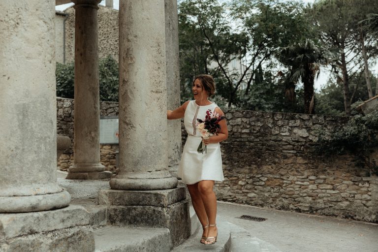 Mariée souriante tenant un bouquet et appuyée contre une colonne en pierre.