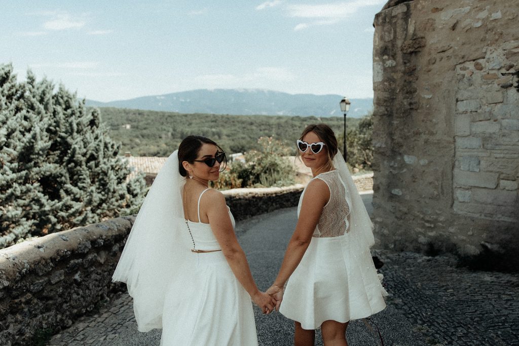 Deux mariées main dans la main regardant par-dessus leurs épaules, l'une avec des lunettes en forme de cœur.