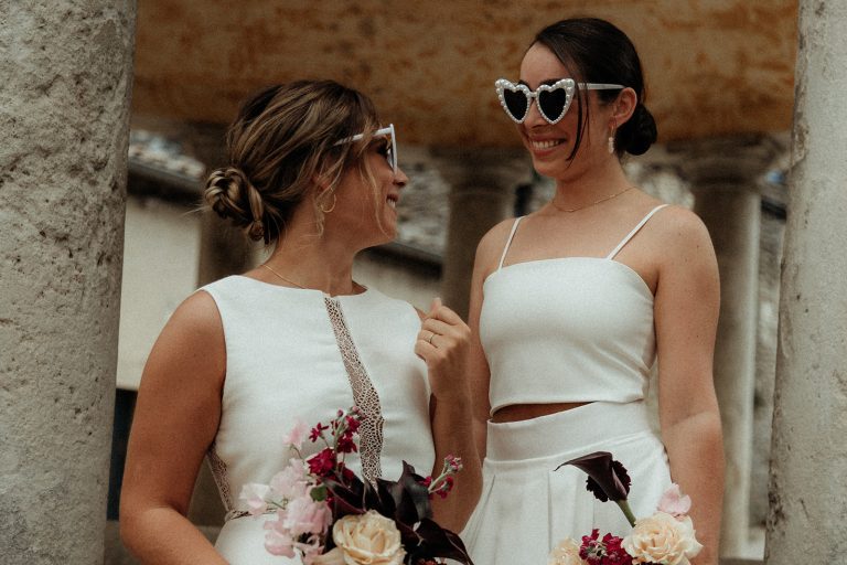 Deux mariées souriantes avec des fleurs, l'une portant des lunettes en forme de cœur.