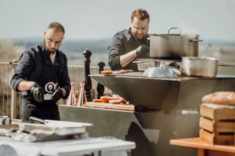 Deux traiteurs en tenue professionnelle cuisinent en plein air sur un brasero avec vue sur la campagne.