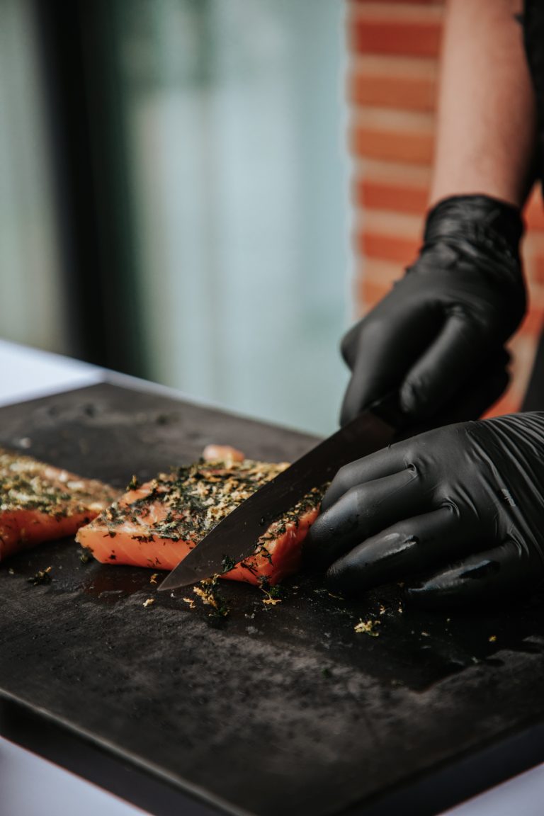 Gros plan sur un chef en train de trancher du saumon frais aux herbes, avec des gants noirs, sur une planche noire.