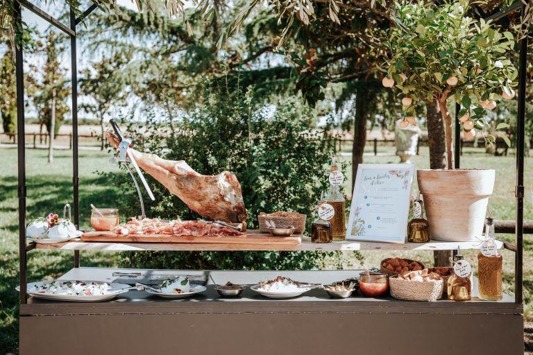 Buffet de mariage en extérieur avec jambon cru à la découpe, huiles aromatisées, pains et accompagnements variés sous un arbre.