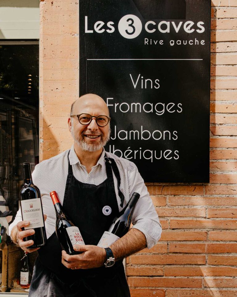 Portrait d’un caviste souriant devant l’enseigne Les 3 Caves Rive Gauche, tenant trois bouteilles de vin dans ses mains.