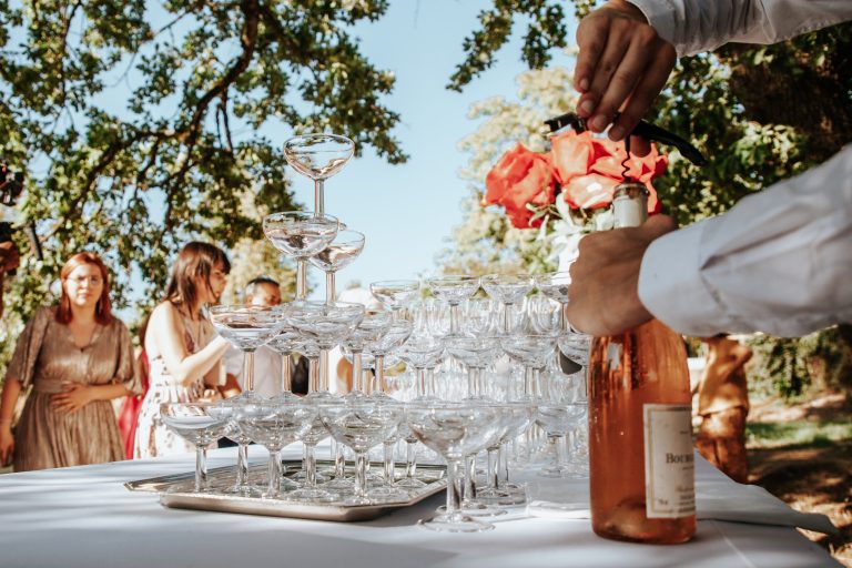 Serveur ouvrant une bouteille de champagne rosé devant une fontaine de coupes pour le vin d’honneur d’un mariage en plein air. Prix traiteur mariage
