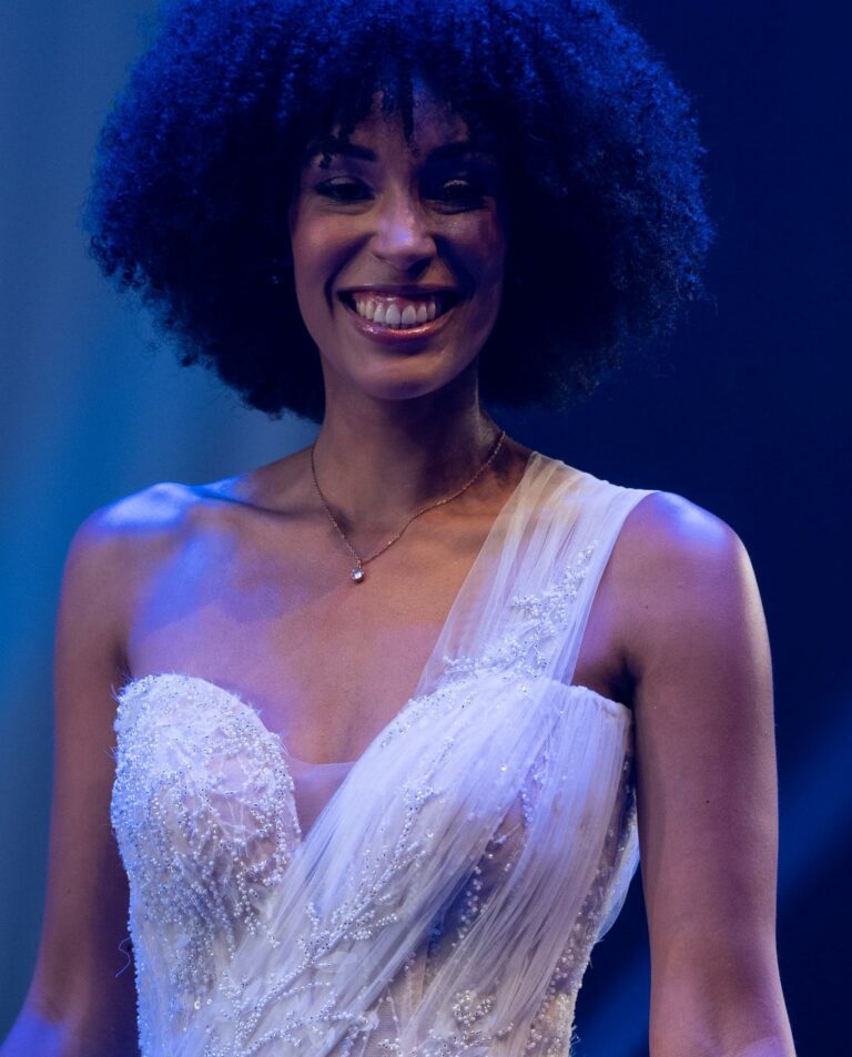 Mariée souriante portant un collier discret avec robe asymétrique brodée.