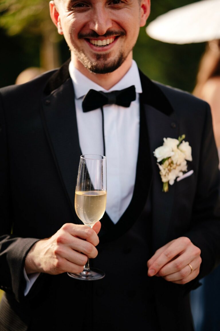 Marié souriant en smoking noir, tenant une flûte de champagne, boutonnière blanche épinglée sur sa veste.