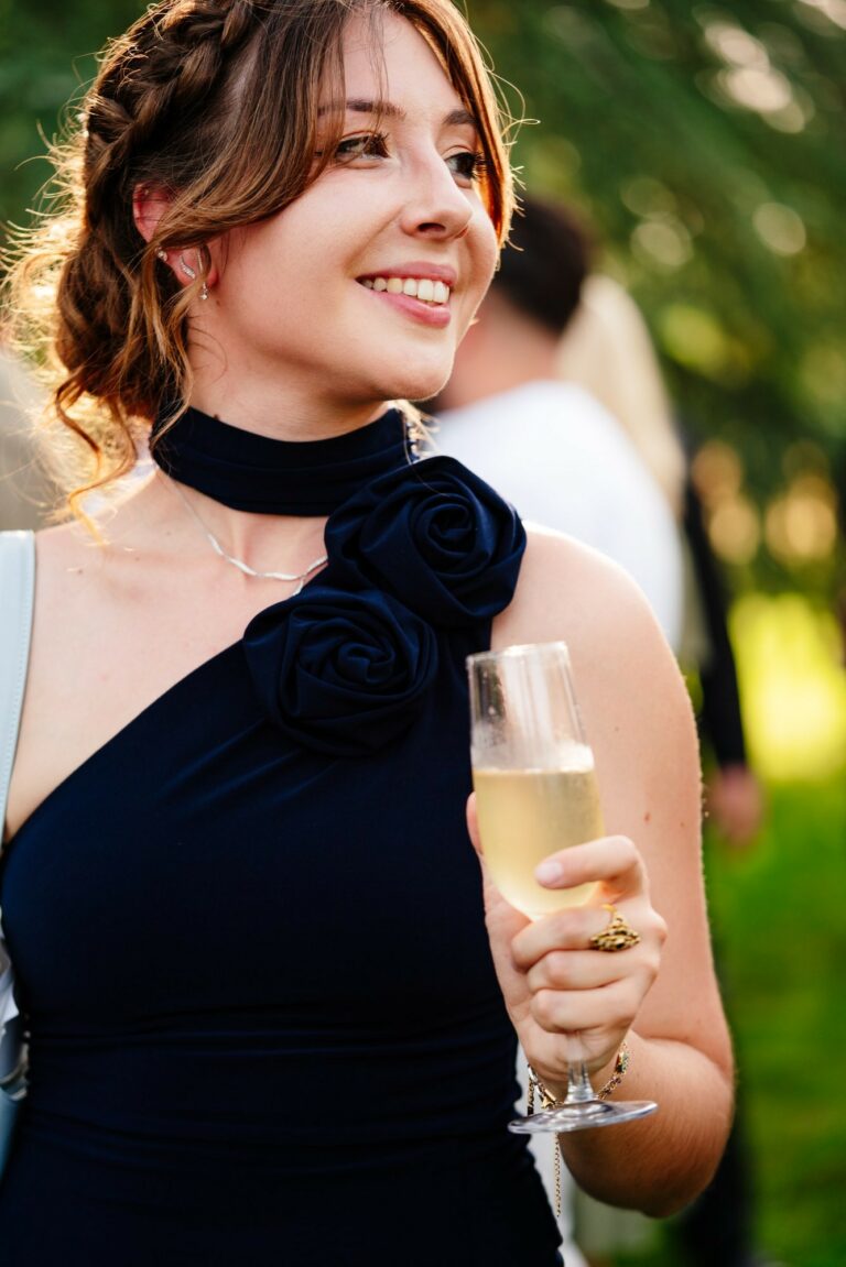 Jeune femme souriante en robe bleu marine ornée de fleurs en tissu, tenant une flûte de champagne lors d’un cocktail de mariage.