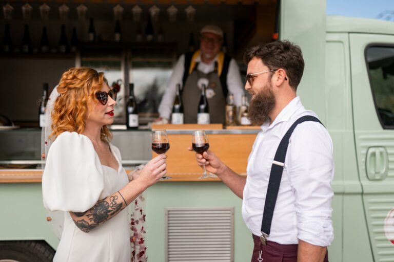 Un couple de jeunes mariés trinque avec des verres de vin rouge devant un food truck vinicole, dans une ambiance rétro et décontractée.