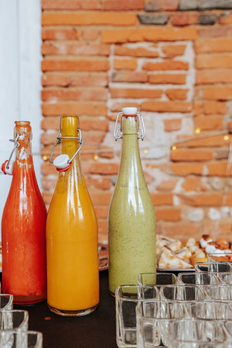 Trois grandes bouteilles de jus faits maison aux couleurs vives – rouge, orange et vert – alignées sur une table de réception avec des verres en fond.