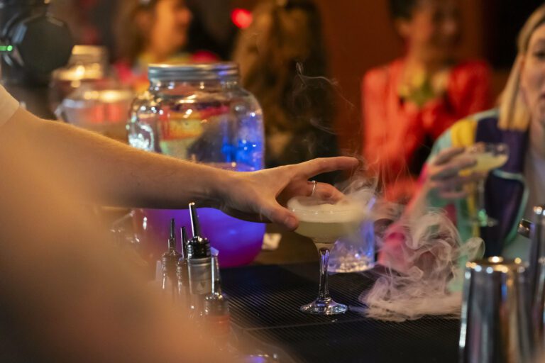 Main d’un barman posant un cocktail fumant sur le comptoir, entouré d’accessoires de mixologie et de convives en arrière-plan.