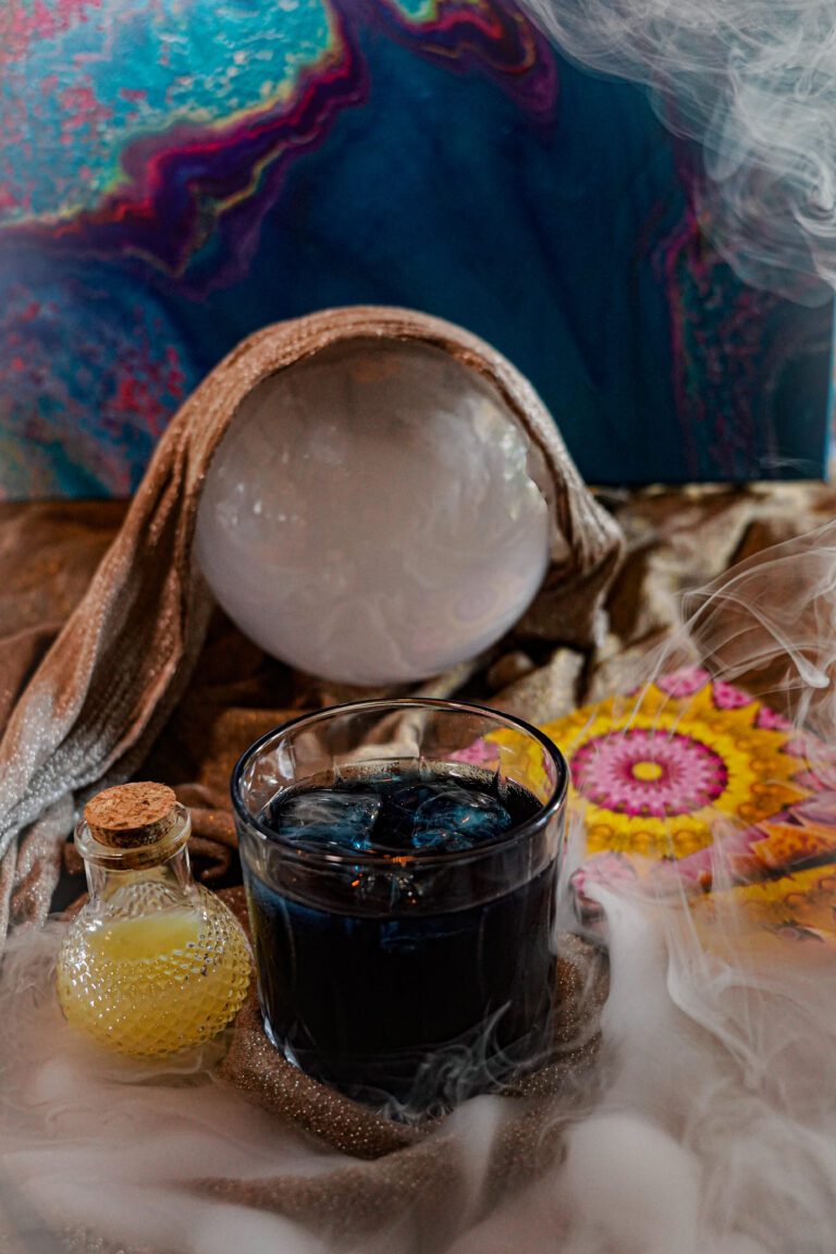 Cocktail bleu nuit avec glaçons, posé devant une boule de cristal et une fiole ancienne, dans une ambiance mystérieuse et brumeuse.