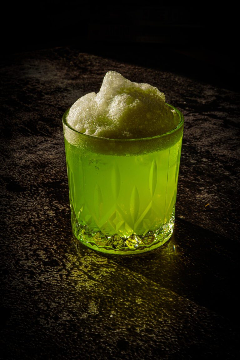 Cocktail vert fluo dans un verre taillé, surmonté d’une mousse légère et brillante, éclairé dans une ambiance tamisée.