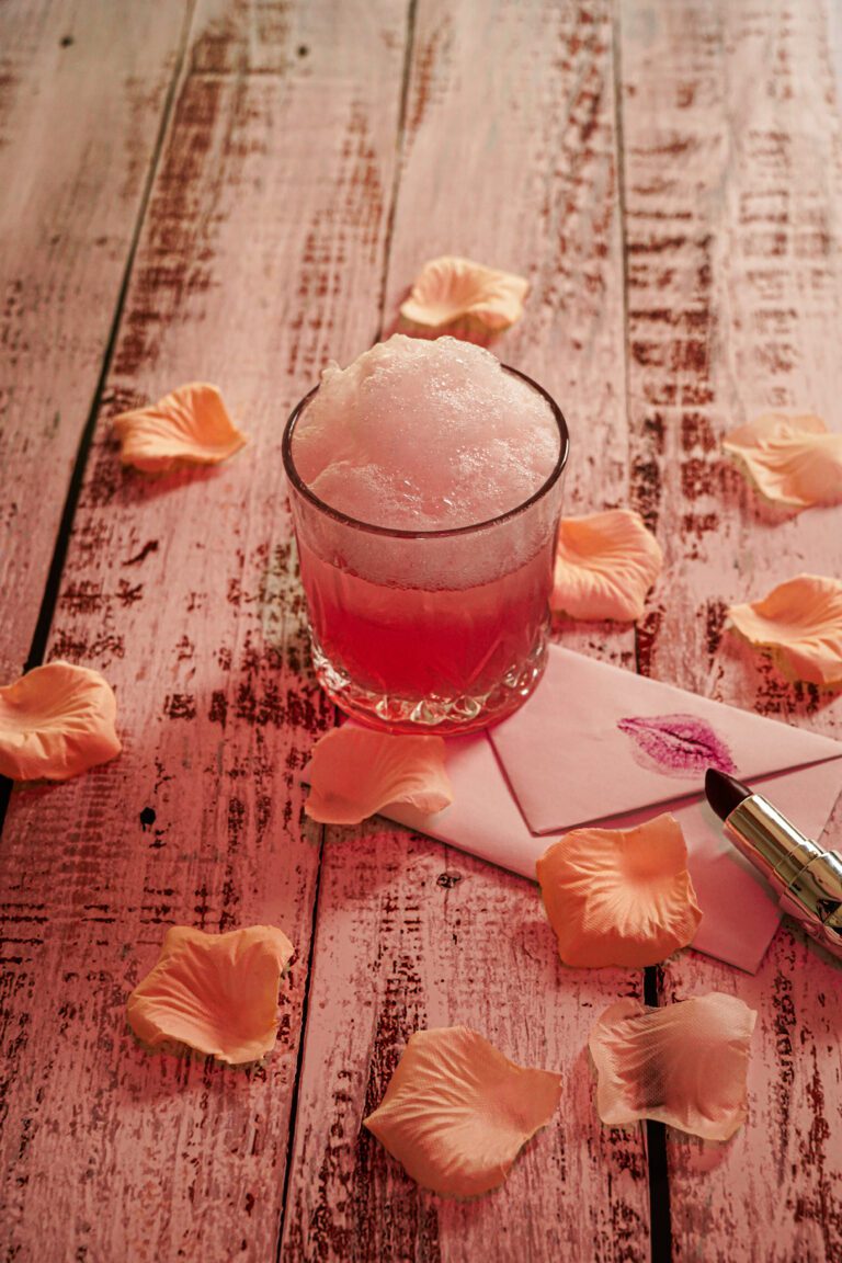 Cocktail rose avec une mousse légère, posé sur une table en bois rose avec des pétales, un rouge à lèvres et une enveloppe marquée d’un baiser.