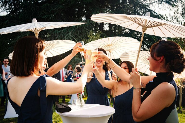 Groupe d’invitées portant des robes bleu marine trinquant avec des coupes de champagne sous des ombrelles blanches pendant un vin d’honneur en extérieur.