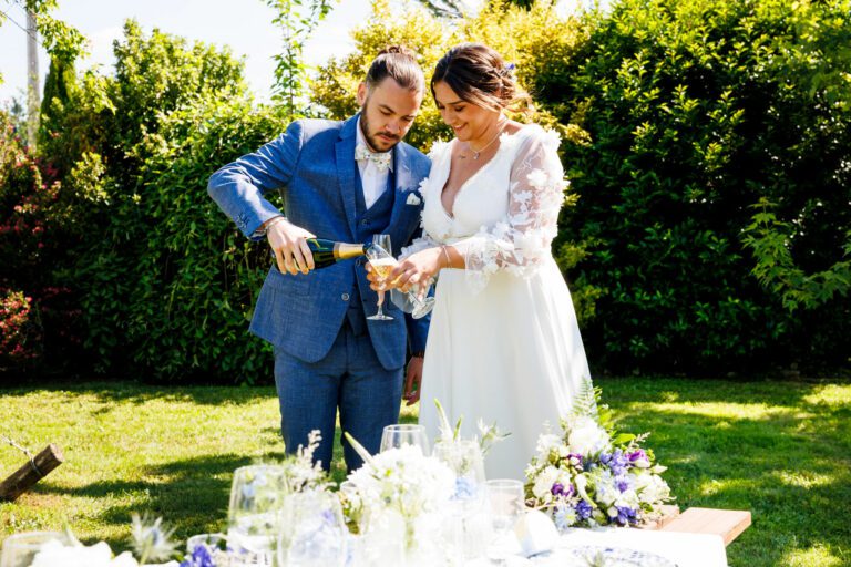 Mariés dans un jardin en train de servir du champagne dans des flûtes, table de réception décorée au premier plan.