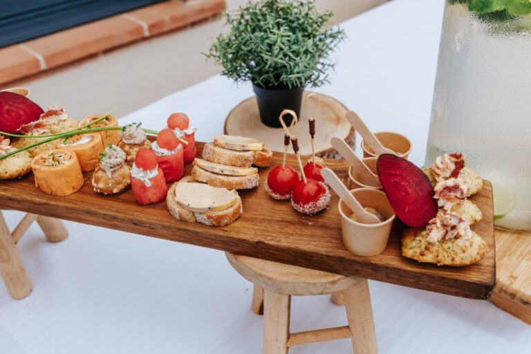 Planche en bois garnie de bouchées apéritives raffinées et colorées : foie gras, billes de pastèque, roulés salés, mini brochettes, verrines et madeleines salées.
