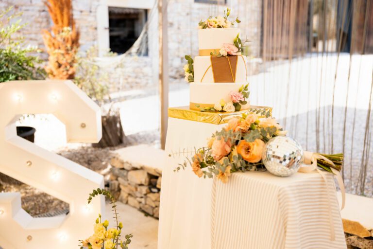 Pièce montée de mariage blanche à trois étages avec ruban doré, ornée de fleurs fraîches et posée sur une table décorée de fleurs, bouquet et boule à facettes.