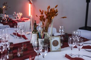 Centre de table de mariage avec bougies torsadées, fleurs séchées, crayons de couleur, menu imprimé sur une bouteille et chemins de table couleur terracotta.