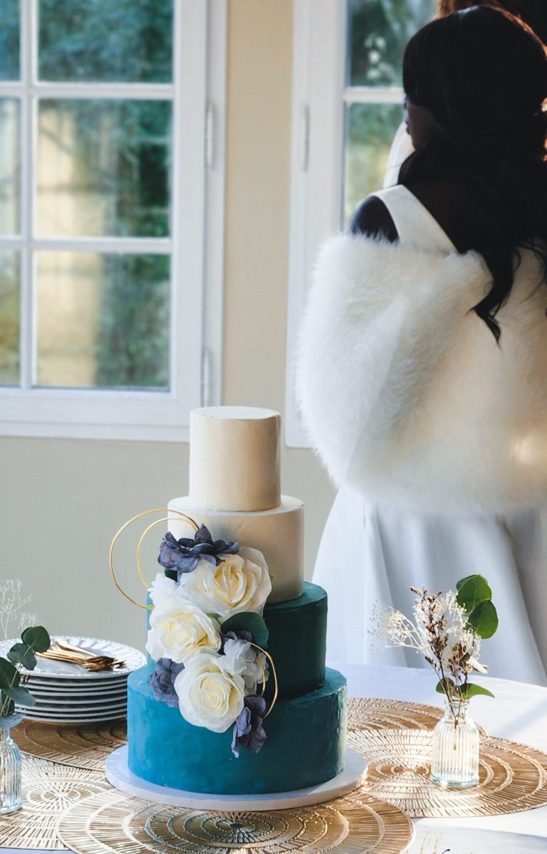 Gros plan sur un wedding cake à trois étages bleu canard et ivoire, décoré de fleurs blanches et violettes, posé sur une table élégante avec sets dorés.