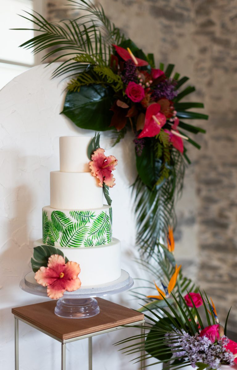 Wedding cake blanc à quatre étages décoré de fleurs d’hibiscus et d’un imprimé feuillage tropical vert.